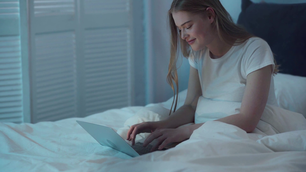 Νεαρή γυναίκα δακτυλογραφεί σε φορητό υπολογιστή και κάθεται στο κρεβάτι  - Πλάνα, βίντεο