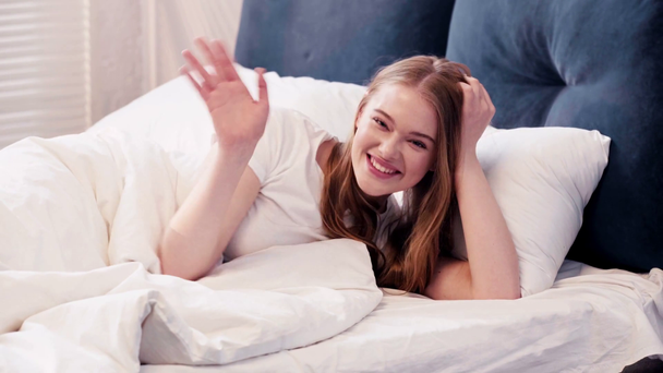 Młoda kobieta leży w łóżku i macha przed kamerą - Materiał filmowy, wideo