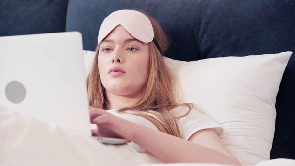 Mujer joven acostada en la cama y escribiendo en el portátil por la mañana - Metraje, vídeo