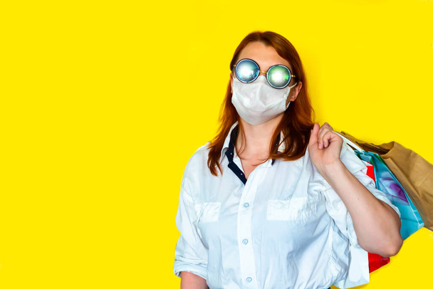 Πορτρέτο του χαρούμενος κορίτσι φορώντας γυαλιά ηλίου και ιατρική μάσκα κρατώντας τσάντες ψώνια ματιά μακριά απομονώνονται σε κίτρινο φόντο, μαύρο Παρασκευή έννοια πώληση σεζόν. - Φωτογραφία, εικόνα