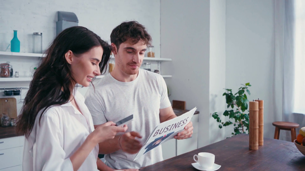 Homem lendo notícias perto de mulher sexy com smartphone e café na cozinha  - Filmagem, Vídeo