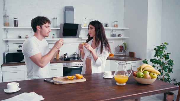 Ο άνθρωπος τρώει πορτοκάλι κοντά σέξι φίλη κατά τη διάρκεια του πρωινού στην κουζίνα  - Πλάνα, βίντεο