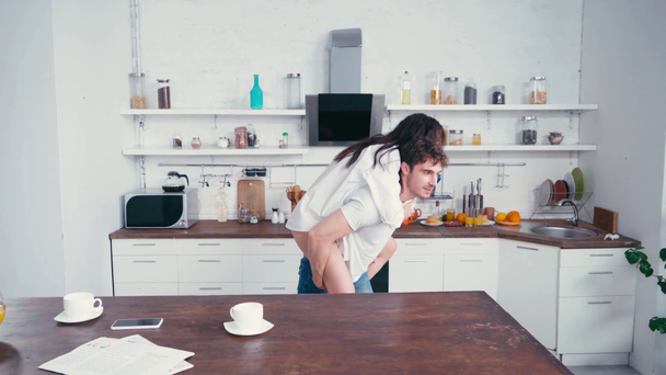 Nainen riggybacking poikaystävä lähellä kahvia ja älypuhelin pöydällä keittiössä  - Materiaali, video