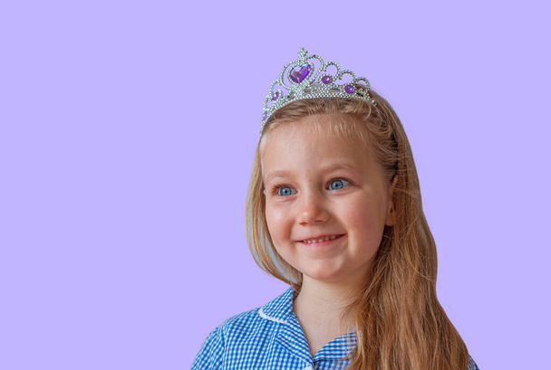 Surprised little princess girl in silver crown looking away with wide open eyes. Open eyed emotional young lady with long wavy hair in blue dress portrait on pastel purple background with copy space. - Photo, Image