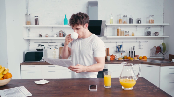 Nuori mies juo kahvia ja lukee uutisia lähellä appelsiinimehua ja älypuhelinta  - Materiaali, video