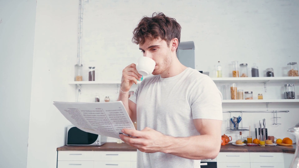 Νεαρός διαβάζει εφημερίδα πίνοντας καφέ στην κουζίνα  - Πλάνα, βίντεο