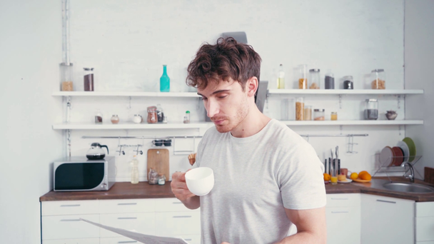 Elinde gazete ve bir fincan kahve olan genç adam mutfakta kameraya bakıyor.  - Video, Çekim