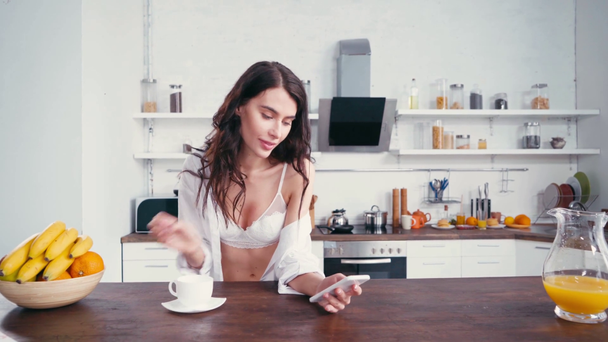 Sütyen ve gömlekli kadın kahve içiyor ve mutfakta akıllı telefon kullanıyor.  - Video, Çekim