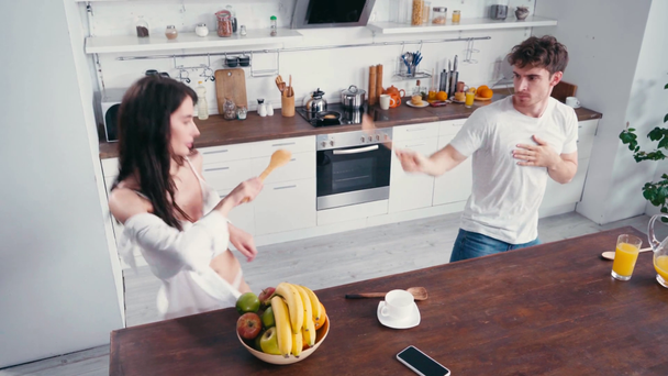 Σέξι ζευγάρι που διασκεδάζουν με σπάτουλες κοντά σε καφέ και φρούτα στην κουζίνα  - Πλάνα, βίντεο