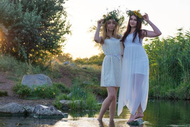 duas namoradas, meninas em vestidos brancos e coroas de flores em suas cabeças, de pé em um rio de água, perto da costa, rindo, contra o pôr do sol. O conceito de virgindade, beleza natural, meninas de vinte anos de pé pés descalços em th - Foto, Imagem