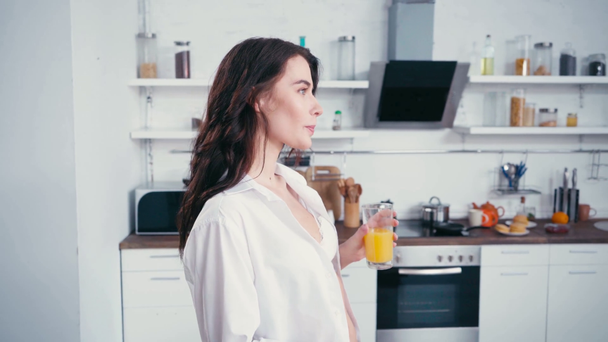 Mujer sexy en camisa bebiendo jugo de naranja en la cocina  - Imágenes, Vídeo