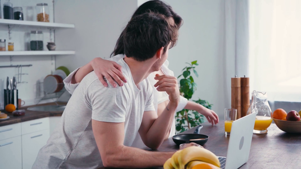 Seductive woman with pancakes in pan embracing man near laptop on table - Video, Çekim