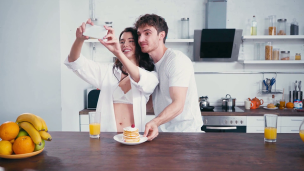 Man with pancakes taking selfie and embracing sexy woman with smartphone at home - Footage, Video