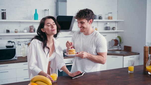 Hombre con panqueques alimentación sexy novia con teléfono inteligente en la cocina  - Imágenes, Vídeo