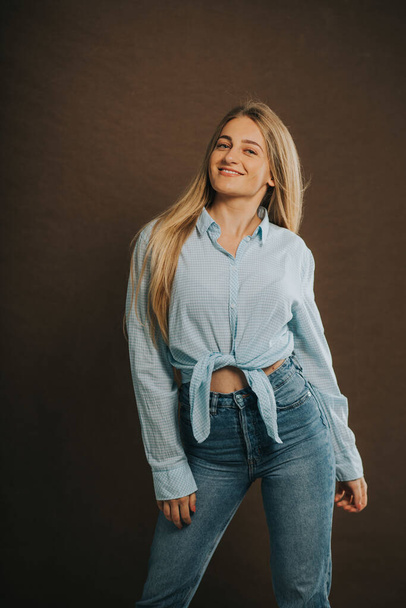 A vertical shot of an attractive blonde female in jeans and a short shirt posing on a brown background - 写真・画像