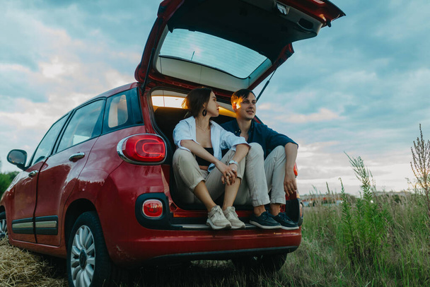 Young couple in love sits and rests inside of opened trunk of red car in meadow. - Photo, Image