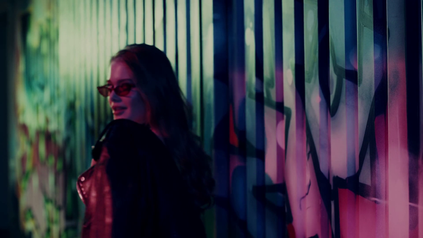 женщина в солнцезащитных очках в кожаной куртке и при ходьбе у стены с граффити - Кадры, видео