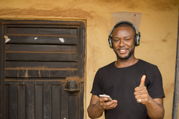 Een jonge Afrikaanse man buiten, die zijn draadloze koptelefoon gebruikt om naar muziek te luisteren op de telefoon. - Foto, afbeelding