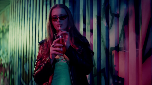 kobieta w okularach przeciwsłonecznych pije koktajl i tańczy przy ścianie z graffiti - Materiał filmowy, wideo