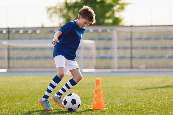 Boy in soccer uniform practice with ball. Child kicking ball on grass stadium. Young athlete improving football dribbling skills on training field. Kid soccer player dribbling through cones - Photo, Image