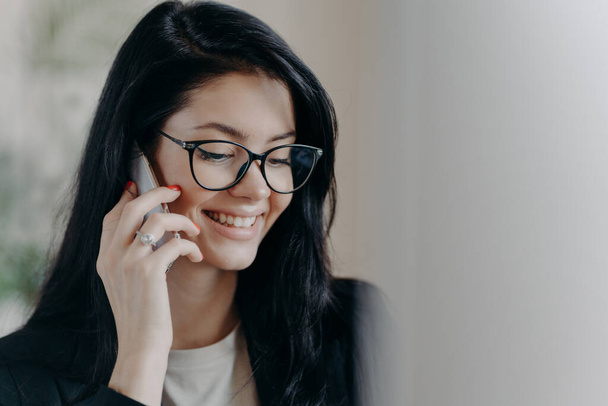 Happy woman lawyer keeps mobile phone near ear, has pleasant conversation with client, discuss working issues, smiles gently, wears transparent glasses, formal wear. Female employee calls someone - Valokuva, kuva