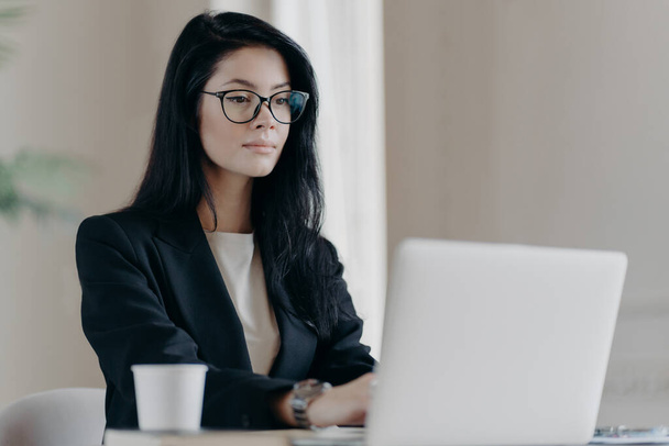 Horizontal shot of concentrated brunette woman sits in front of laptop computer, wears glasses, creats new post on website, sits in coworking space with paper cup of coffee. Manager at work. - Foto, Imagem