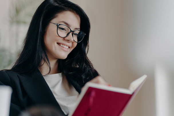 Cheerful dark haired woman creats do list with targets, holds notebook, writes organisation plan, wears spectacles, dressed in formal outfit, smiles pleasantly. Autodidact and education concept - Фото, зображення