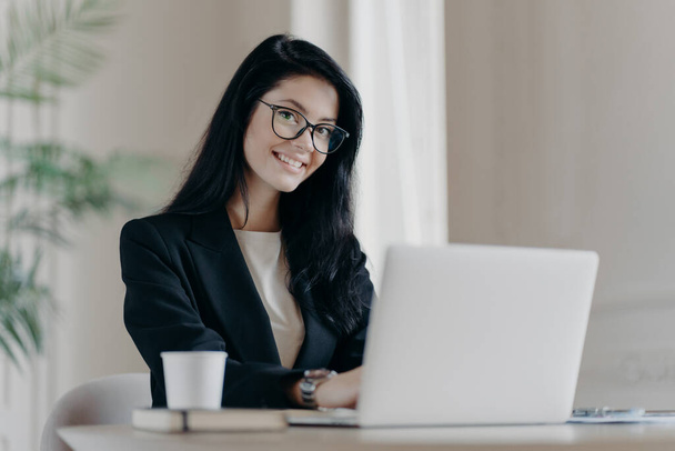 Positive dark haired woman makes research, browses information on laptop computer, connected to wireless internet, being happy wears formal clothes and spectacles drinks coffee. Economist works online - Photo, Image