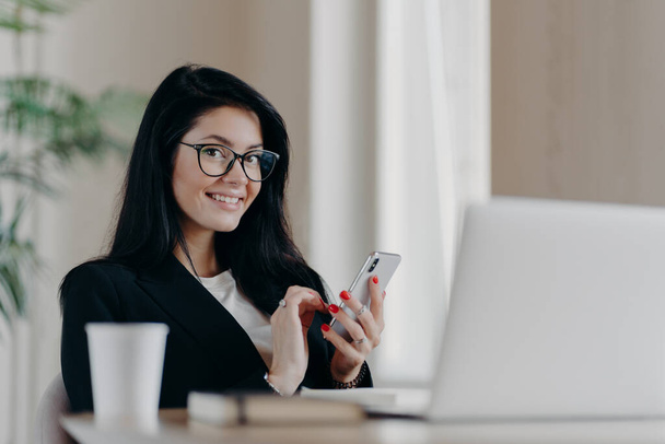 Horizontální záběr šťastné brunetky Evropské ženy vzkazy na moderním smartphonu, poskytuje online konzultaci, usměje se pozitivně, nosí brýle a formální oblek, sedí před otevřeným notebookem. - Fotografie, Obrázek