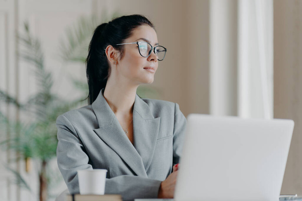 成功したブルネットの女性起業家は、思慮深い表現とは別に集中し、ラップトップコンピュータを介してウェビナーを見るために行く、透明メガネを着用し、正式な服を着て、コーヒーを飲む - 写真・画像