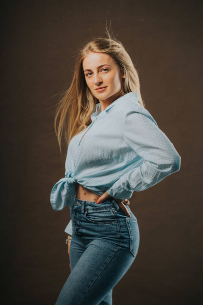A vertical shot of an attractive blonde female in jeans and a short shirt posing on a brown background - Valokuva, kuva