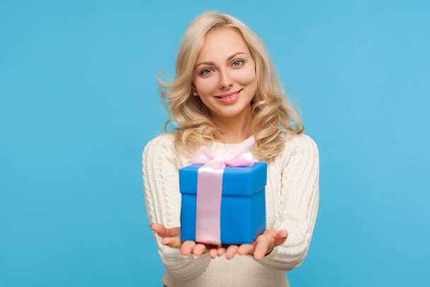 Cute positive blond woman in knitted sweater holding in hands blue giftbox with ribbon bow, making present, surprise. Indoor studio shot isolated on blue background - Photo, Image