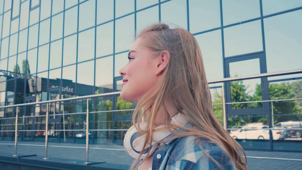 junge Frau mit kabellosen Kopfhörern blickt auf Kamera in der Nähe von Gebäude  - Filmmaterial, Video
