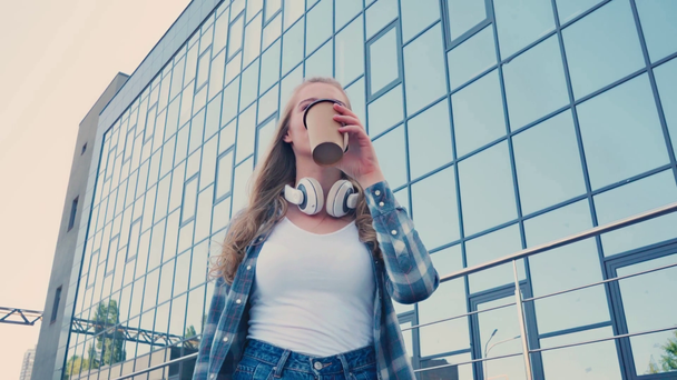 visão de baixo ângulo da mulher bebendo café para ir perto do edifício com fachada de vidro - Filmagem, Vídeo