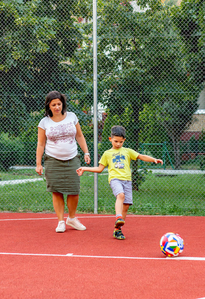 POZNAN, POLSKO - 26. srpna 2020: Mladý šestiletý chlapec kopal do fotbalu v blízkosti ženy na veřejném hřišti - Fotografie, Obrázek