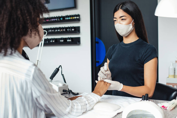 Salão de manicure e pedicure, conceito covid-19. Menina asiática em máscara protetora desinfecta unhas para o cliente - Foto, Imagem