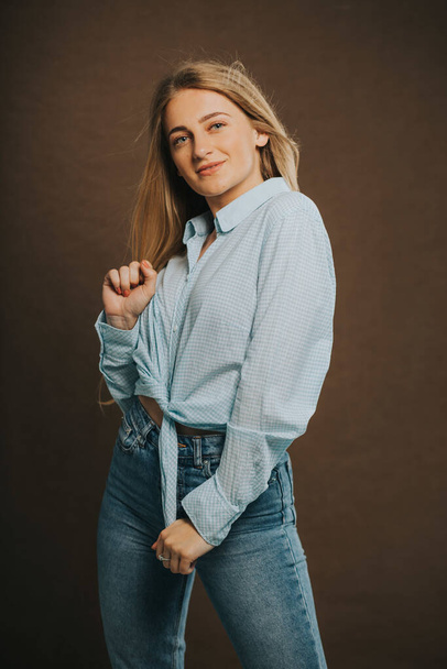 A vertical shot of an attractive blonde female in jeans and a short shirt posing on a brown background - Фото, изображение
