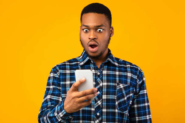 Σοκαρισμένος μαύρος κοιτάζοντας Smartphone ανάγνωση SMS, κίτρινο φόντο - Φωτογραφία, εικόνα