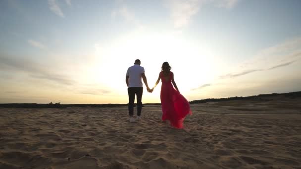 Kamera sledující mladé páry, držící se za ruce a procházející se pouští. Pár milenců na písku při západu slunce. Koncept letní cesty nebo dovolené. Scénická krajina se slunečním světlem - Záběry, video