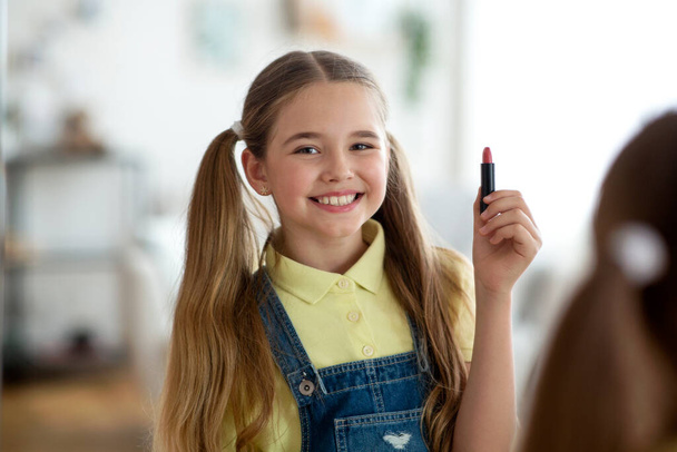 Joven chica sosteniendo lápiz labial, mirando el espejo en el dormitorio - Foto, imagen