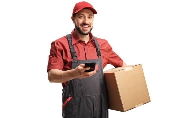 Man courier holding a cardboard box and a mobile phone looking at the camera isolated on white background  - Zdjęcie, obraz