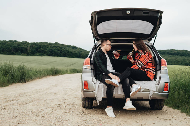 Νεαρό όμορφο ζευγάρι κάθεται στο αυτοκίνητο Trunk και απολαμβάνοντας το οδικό ταξίδι, Picnic Concept - Φωτογραφία, εικόνα