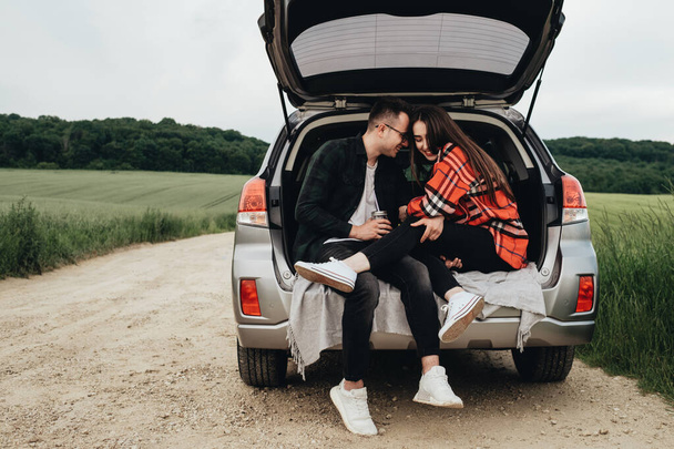 Young Beautiful Couple Sitting in Car Trunk and Enjoying the Roadtrip - Photo, Image