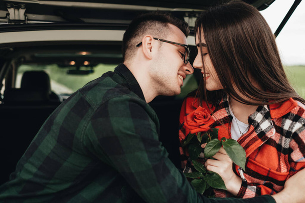 Νεαρό όμορφο ζευγάρι κάθεται στο αυτοκίνητο Trunk και απολαμβάνοντας την Roadtrip, κορίτσι κρατώντας ένα τριαντάφυλλο, ρομαντική διάθεση - Φωτογραφία, εικόνα