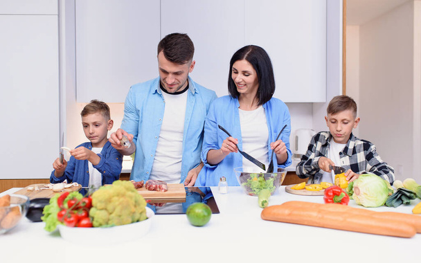 Mom, dad and two sons preparing together healthy breakfast with meat and vegetables on cozy kitchen at dining table. - Фото, изображение