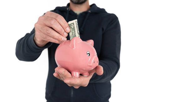 Mann hält Sparschwein in der einen Hand und steckt mit der anderen Dollarschein hinein - Foto, Bild