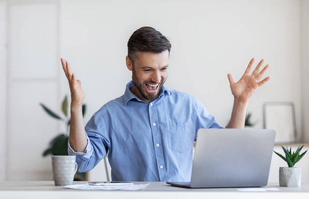 Happy male entrepreneur emotionally celebrating business success with laptop in office - Foto, Imagen