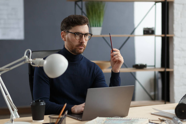 Architect working in office with laptop.Engineer thinks over architectural plan, searching new ideas for construction project. Portrait of handsome bearded man sitting at workplace. Business concept - Foto, Imagem