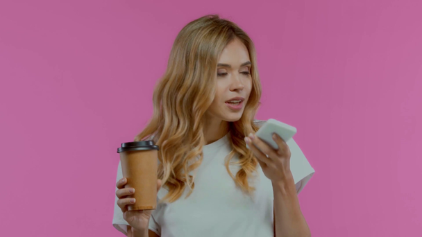 Mujer rubia usando teléfono inteligente y beber café para ir aislado en púrpura - Imágenes, Vídeo