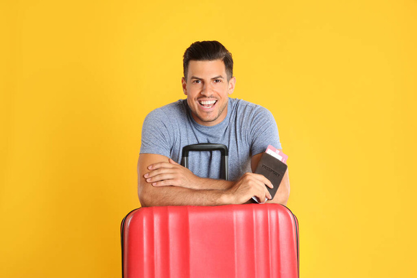 Handsome man with suitcase and ticket in passport for summer trip on yellow background. Vacation travel - Valokuva, kuva